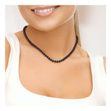 Une femme vêtue d'un haut blanc sans manches sourit doucement, portant le Perlinea Collier NOELYSE - un élégant collier en argent orné de perles noires de Tahiti de 6/7 mm et d'un fermoir Boule.