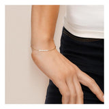 Un gros plan montre un poignet portant le bracelet Perlinea NANÉA | Or Jaune & Perles Blanches, un délicat bracelet en or avec cinq perles blanches, associé à un haut blanc et un pantalon noir. La main détendue repose naturellement à ses côtés.