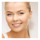 Une jeune femme blonde à la peau claire et aux yeux bleus sourit à l'appareil photo, portant Perlinea Boucle d'Oreilles Bouton AYA en argent 925 et perles de culture blanches 4/5mm sur un fond clair et neutre.