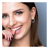 Une femme aux longs cheveux bruns et aux yeux clairs sourit, portant des Boucles d'Oreilles Demi Créole 25 mm Oxydes de Zirconium Beloved et du vernis à ongles noir. Elle pose contre un mur gris uni, la main près de la bouche.