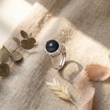 Bague argent 925 ornée d'une perle noire de Tahiti entourée de zircons, posée sur tissu beige.