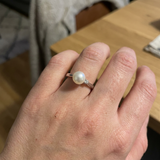 Bague en or blanc 9 carats portée à la main, ornée d'une perle blanche ronde et d'un petit diamant.