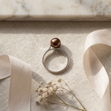 Bague or blanc 9 carats sertie d'une perle de culture chocolat, posée sur tissu beige.