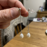 Boucles d'oreilles pendantes or blanc 9 carats avec chaîne fine et perles blanches, tenues à la main.