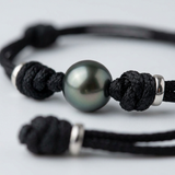 Gros plan sur la perle de Tahiti d'un bracelet homme en coton ciré noir avec nœuds et anneaux argentés.