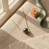 Collier en or jaune 9 carats avec une perle de Tahiti grise posé sur un tissu beige.